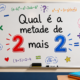 Imagem de uma sala de aula sem pessoas, com um quadro branco exibindo a pergunta “Qual é a metade de 2 mais 2?”, números 2 e 3 em destaque, vários símbolos matemáticos coloridos, apagador e marcador na parte inferior.