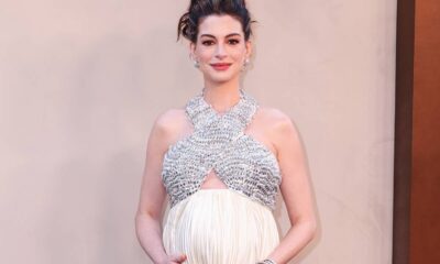 Anne Hathaway, visivelmente grávida, vestindo top brilhante prateado e saia branca, sorrindo