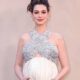 Anne Hathaway, visivelmente grávida, vestindo top brilhante prateado e saia branca, sorrindo