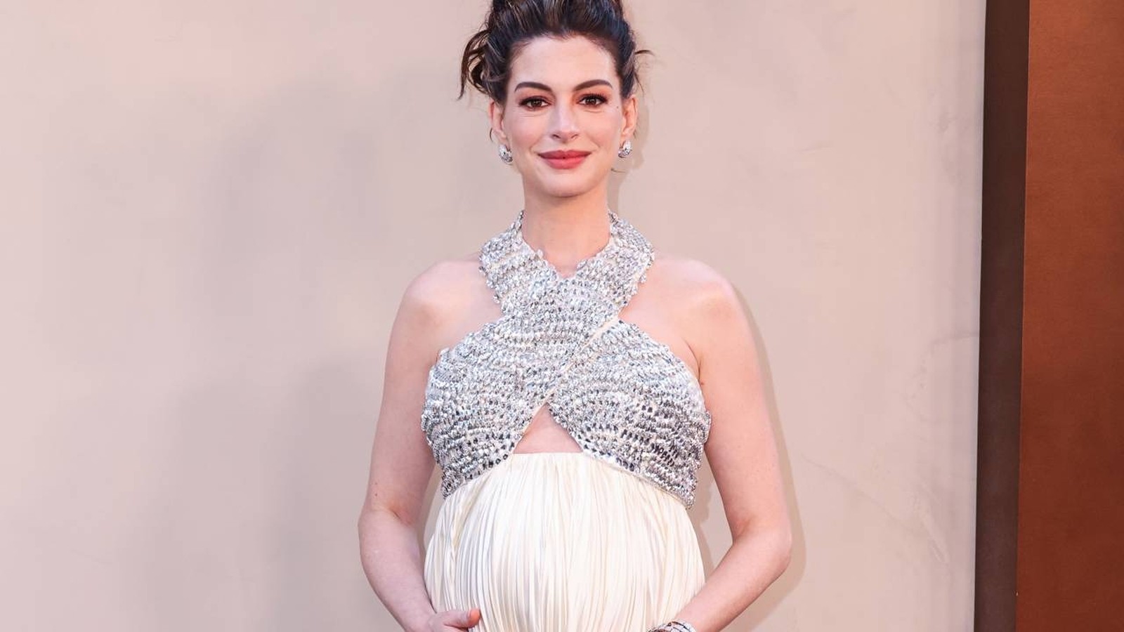Anne Hathaway, visivelmente grávida, vestindo top brilhante prateado e saia branca, sorrindo