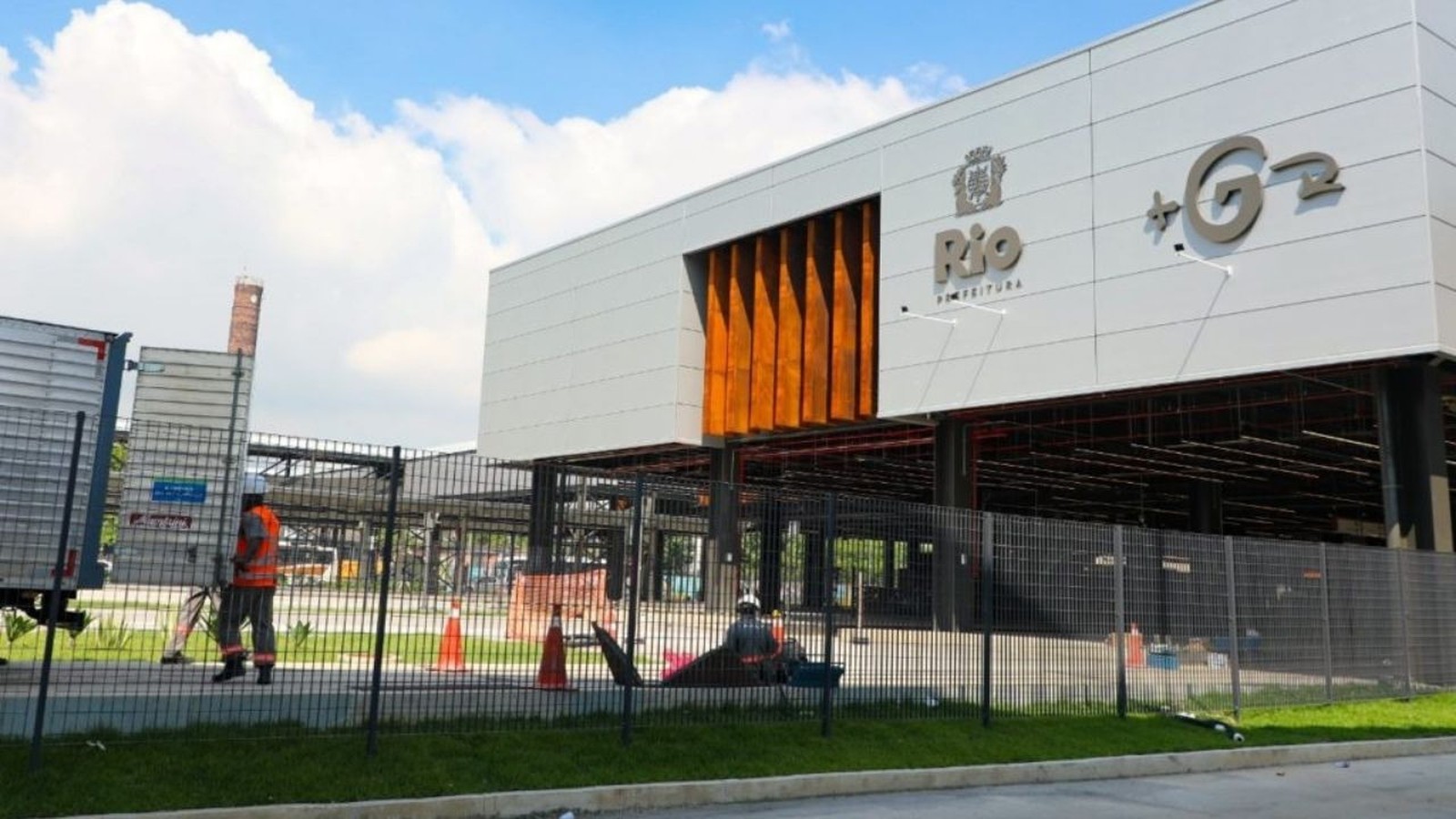Edifício moderno branco com logo 'Rio Prefeitura' e G2, cerca, trabalhador e caminhão à frente