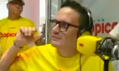 Homem de óculos, fones amarelos e camiseta "tropicana" gesticula em estúdio de rádio
