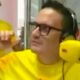Homem de óculos, fones amarelos e camiseta "tropicana" gesticula em estúdio de rádio