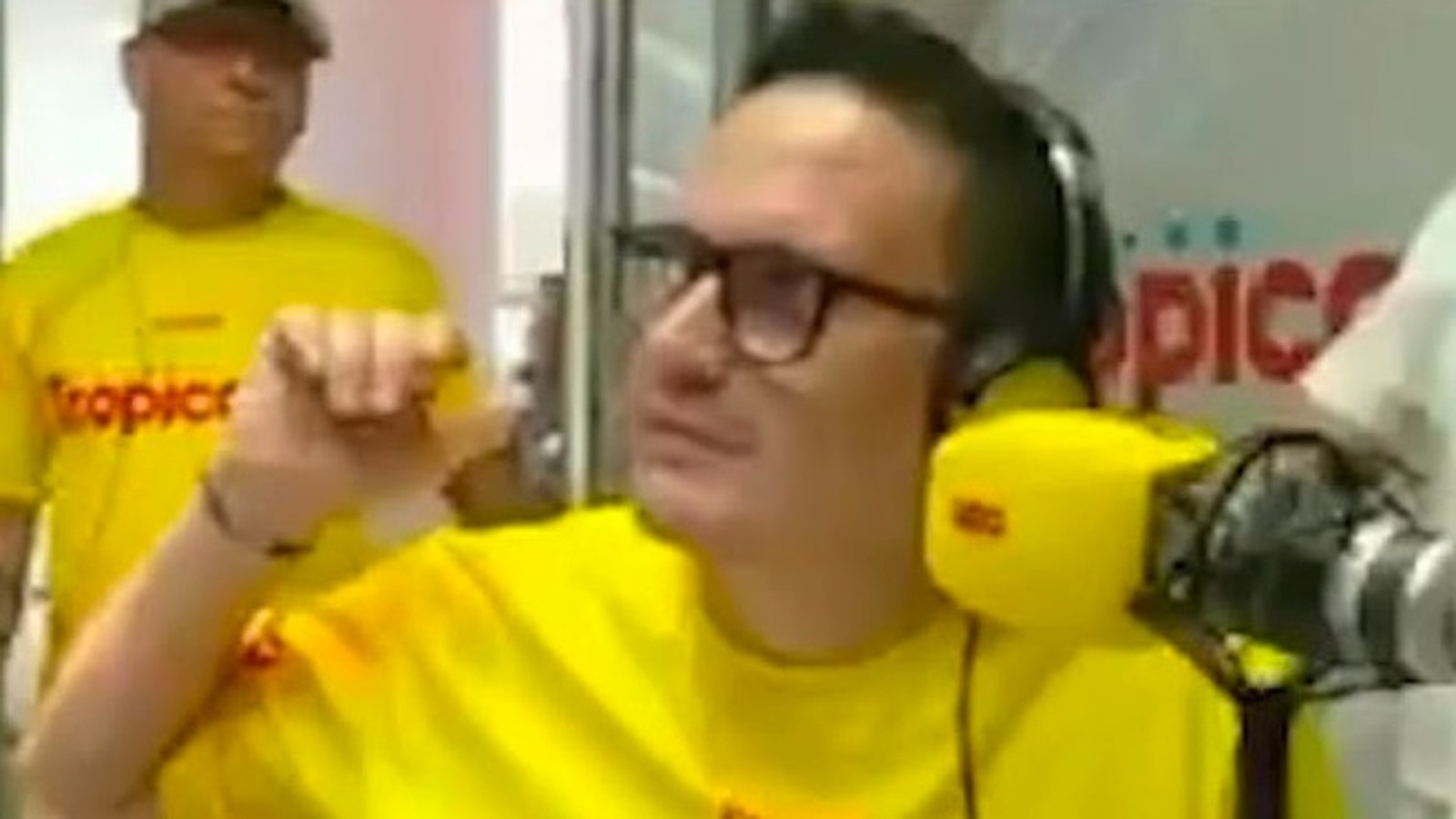Homem de óculos, fones amarelos e camiseta "tropicana" gesticula em estúdio de rádio