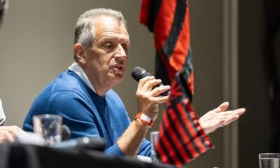 Homem sênior de camisa azul falando em um microfone e gesticulando, com bandeira rubro-negra atrás