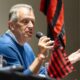 Homem sênior de camisa azul falando em um microfone e gesticulando, com bandeira rubro-negra atrás