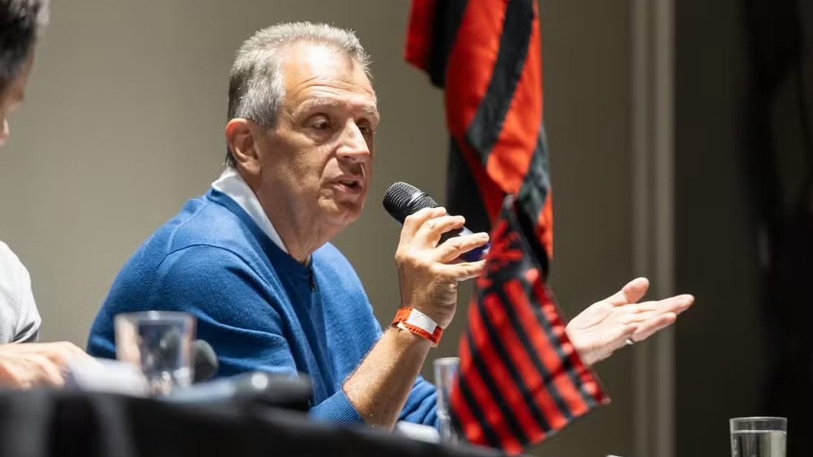 Homem sênior de camisa azul falando em um microfone e gesticulando, com bandeira rubro-negra atrás