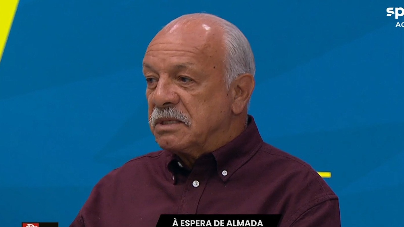 Homem idoso, bigode grisalho, camisa bordô, fundo azul. Logos "SPORTV" e texto "À ESPERA DE ALMADA"