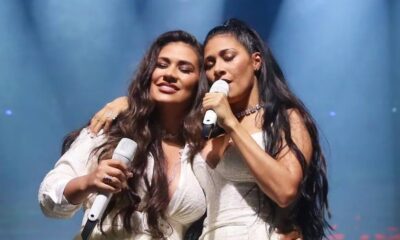 Duas mulheres com microfones cantando no palco, uma sorrindo e a outra de olhos fechados