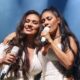 Duas mulheres com microfones cantando no palco, uma sorrindo e a outra de olhos fechados