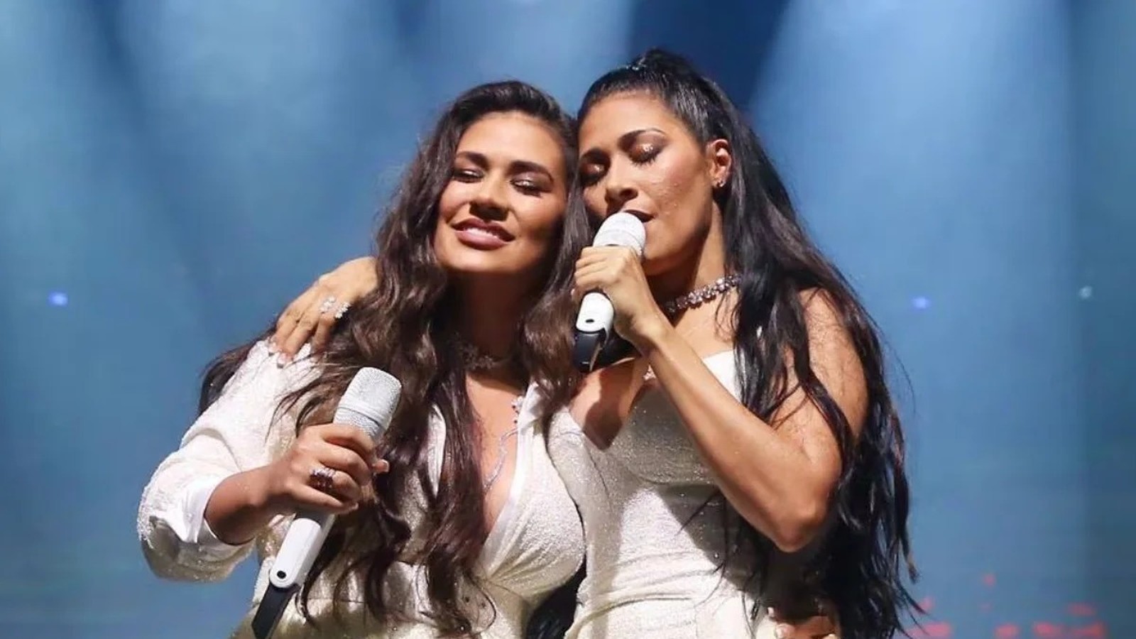 Duas mulheres com microfones cantando no palco, uma sorrindo e a outra de olhos fechados