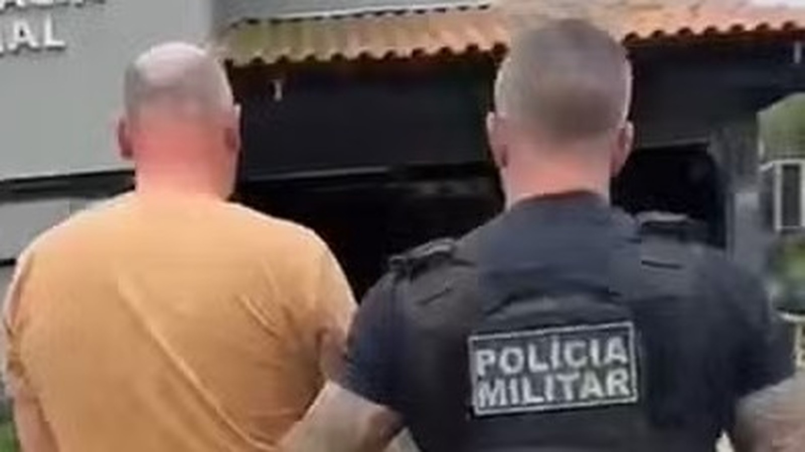 Policial militar com 'POLICIA MILITAR' na camisa escolta homem em camiseta laranja