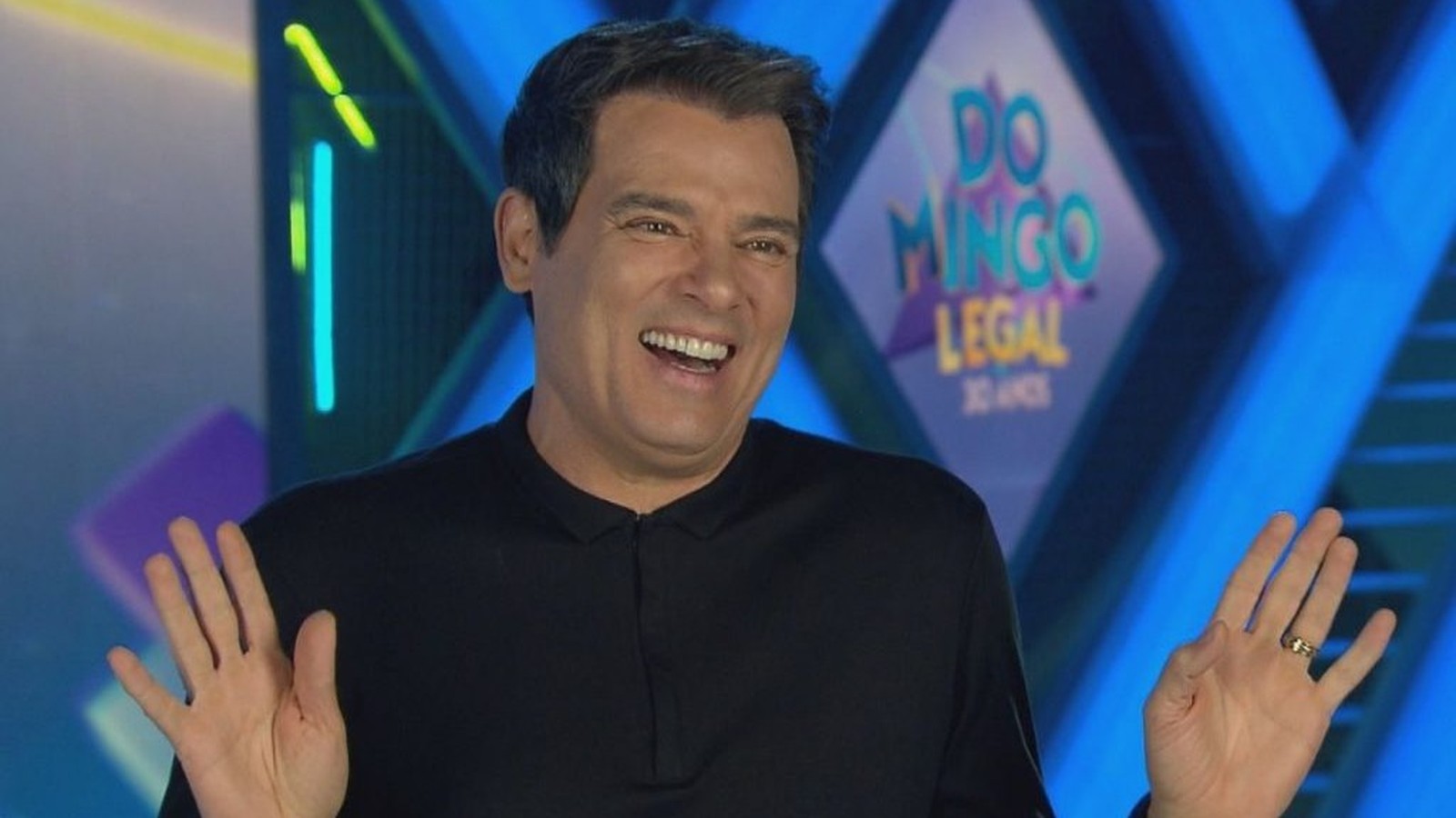 Celso Portiolli sorrindo e gesticulando com logo Domingo Legal no fundo