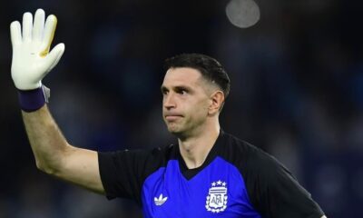 Dibu Martínez com camisa da Argentina e luva branca de goleiro, braço levantado
