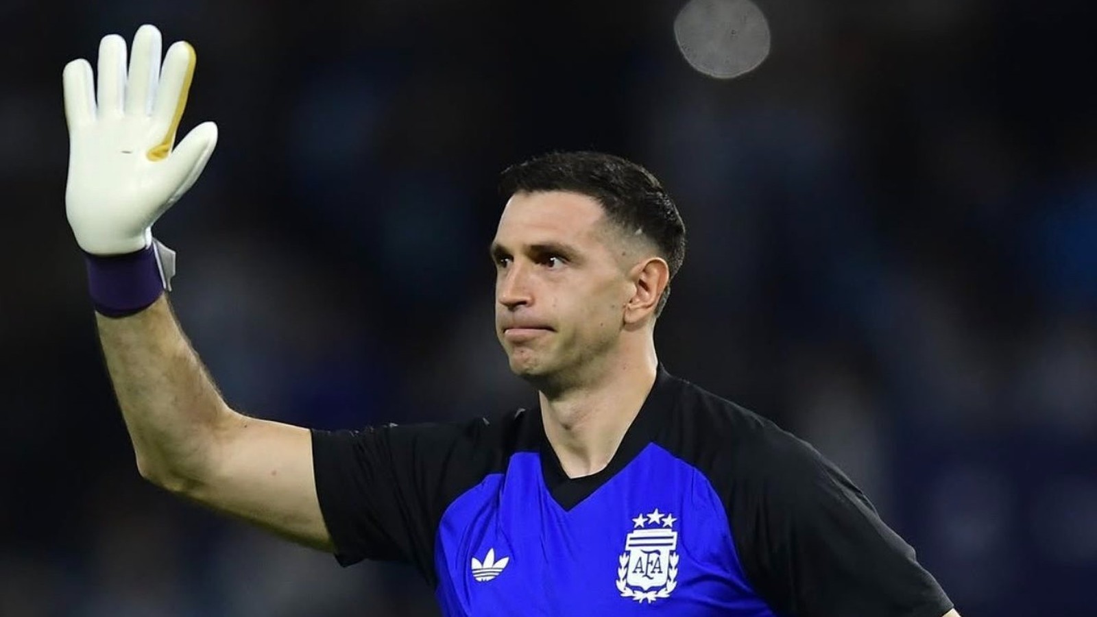 Dibu Martínez com camisa da Argentina e luva branca de goleiro, braço levantado