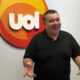 Homem sorridente gesticula à frente de logo UOL branco com círculos coloridos
