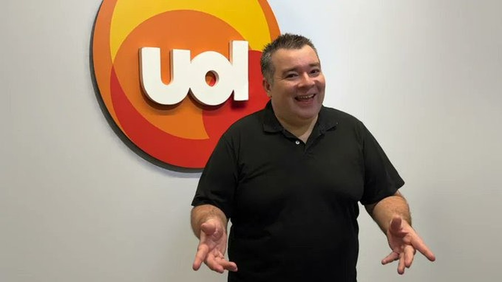 Homem sorridente gesticula à frente de logo UOL branco com círculos coloridos