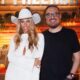 Rodrigo Carelli com óculos e mulher loira de chapéu de cowboy branco em cenário rústico