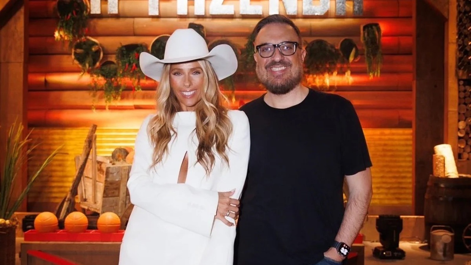 Rodrigo Carelli com óculos e mulher loira de chapéu de cowboy branco em cenário rústico