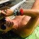 Mulher deitada de costas sem roupa em uma toalha verde, chapéu e óculos escuros
