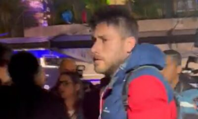 Homem de perfil com jaqueta azul e vermelha, barba, entre pessoas em ambiente noturno