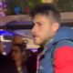 Homem de perfil com jaqueta azul e vermelha, barba, entre pessoas em ambiente noturno