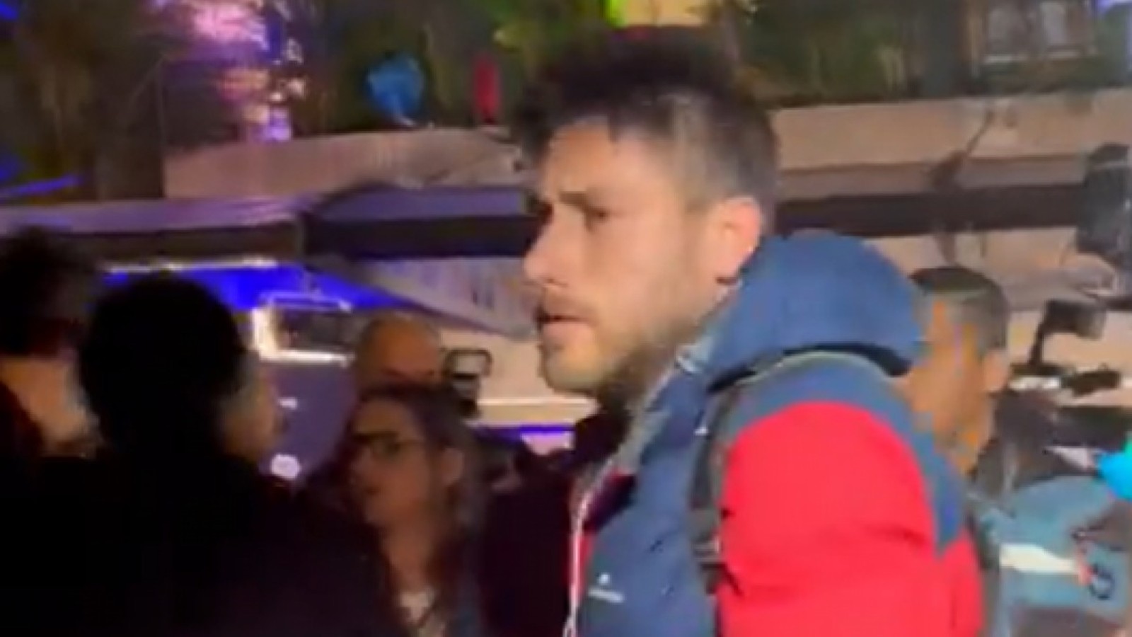 Homem de perfil com jaqueta azul e vermelha, barba, entre pessoas em ambiente noturno