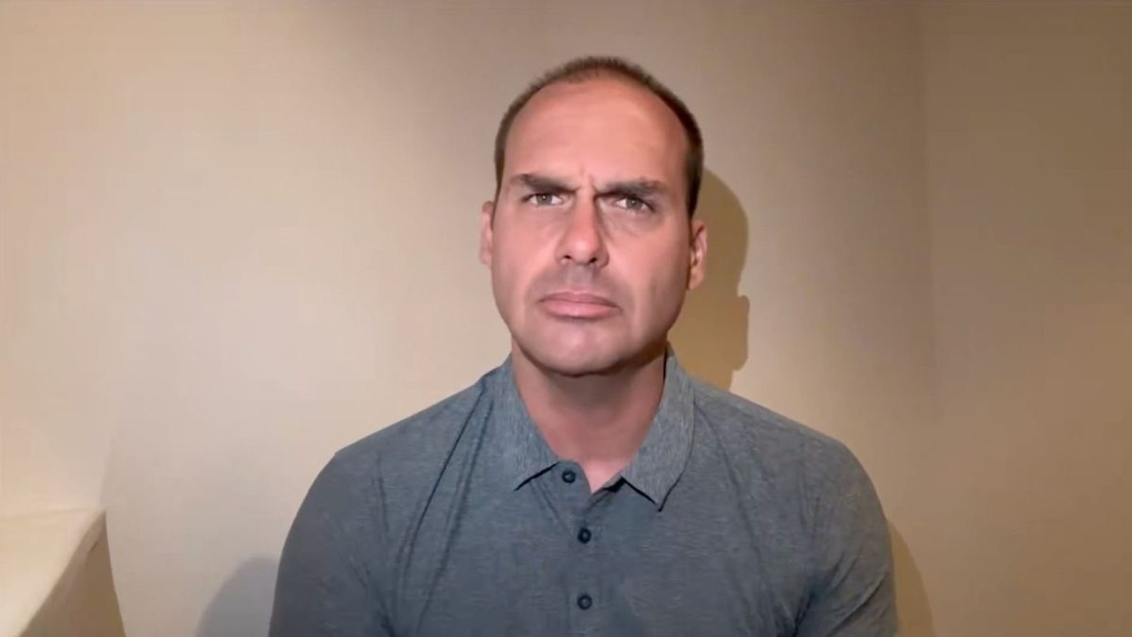 Homem de meia-idade careca, testa franzida, camisa polo cinza, contra parede bege