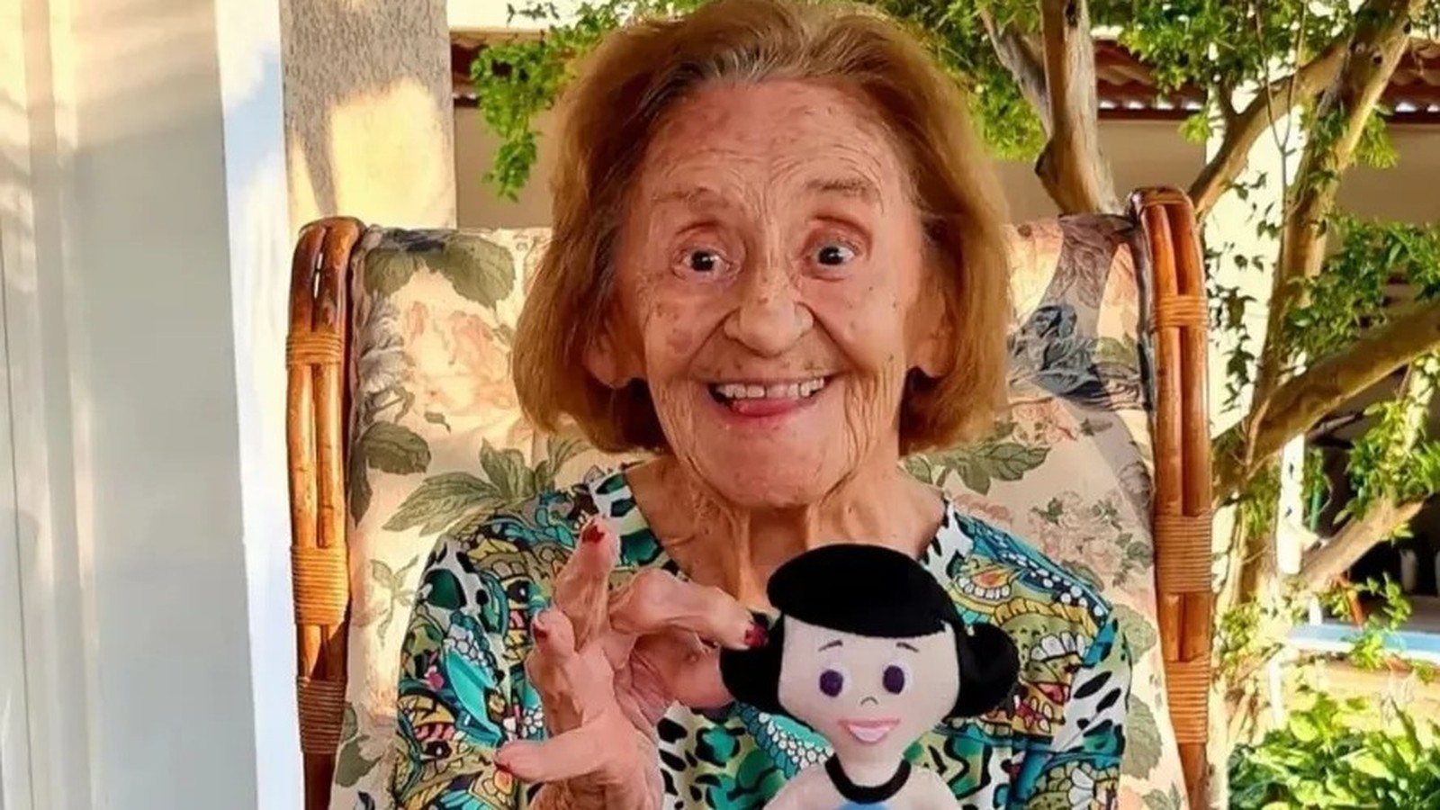 Mulher idosa sorrindo, cabelo castanho-avermelhado, sentada em cadeira, segurando boneca Mônica