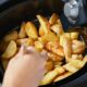 Mão de pessoa mexendo batatas rústicas douradas dentro do cesto de uma airfryer preta