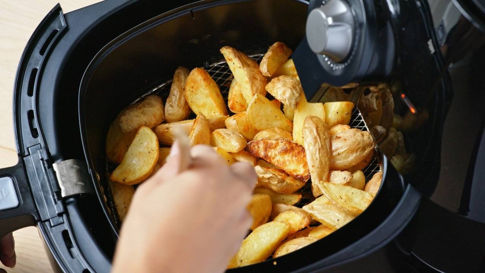 Mão de pessoa mexendo batatas rústicas douradas dentro do cesto de uma airfryer preta