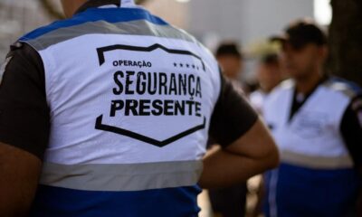 Costas de um agente de segurança com colete branco e azul escrito "OPERAÇÃO SEGURANÇA PRESENTE"