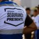 Costas de um agente de segurança com colete branco e azul escrito "OPERAÇÃO SEGURANÇA PRESENTE"