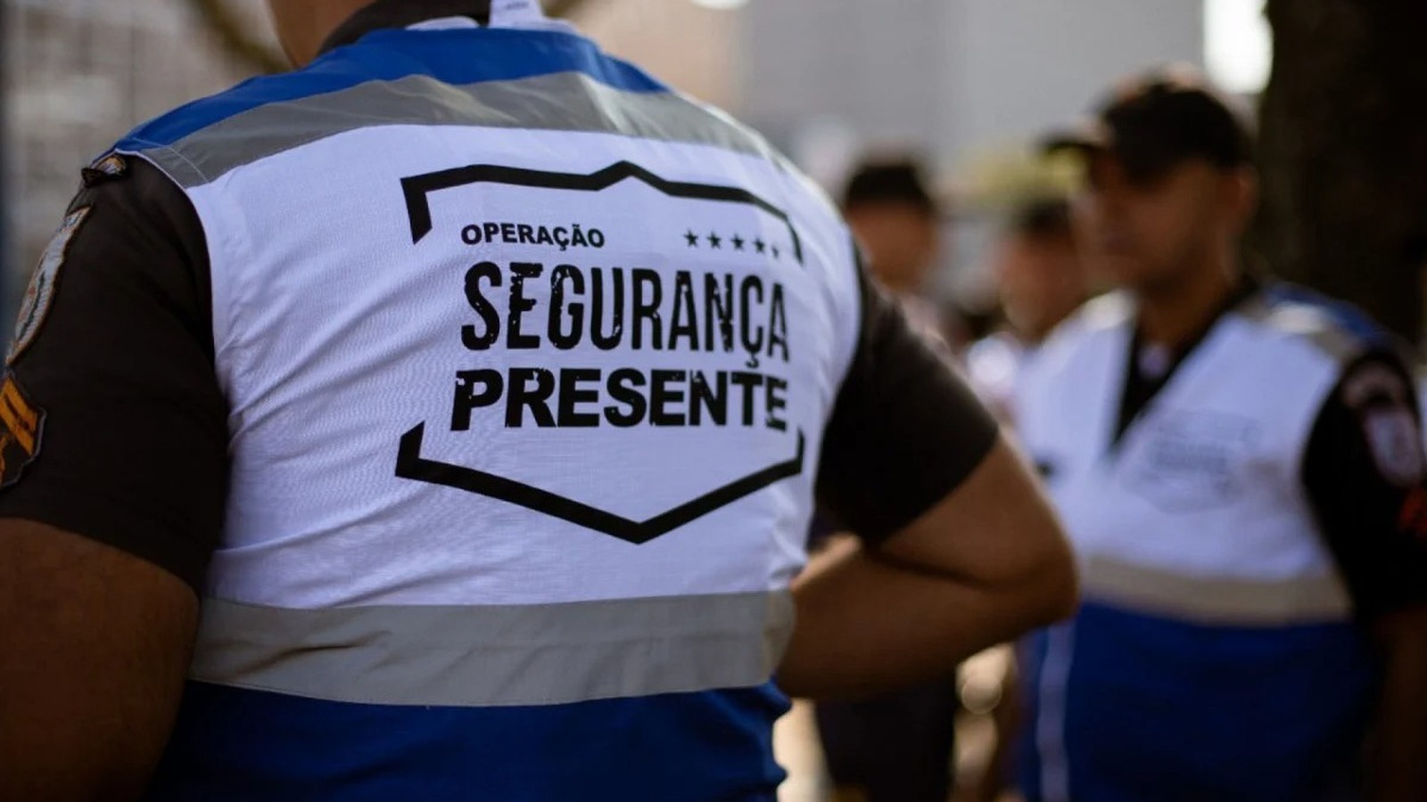 Costas de um agente de segurança com colete branco e azul escrito "OPERAÇÃO SEGURANÇA PRESENTE"