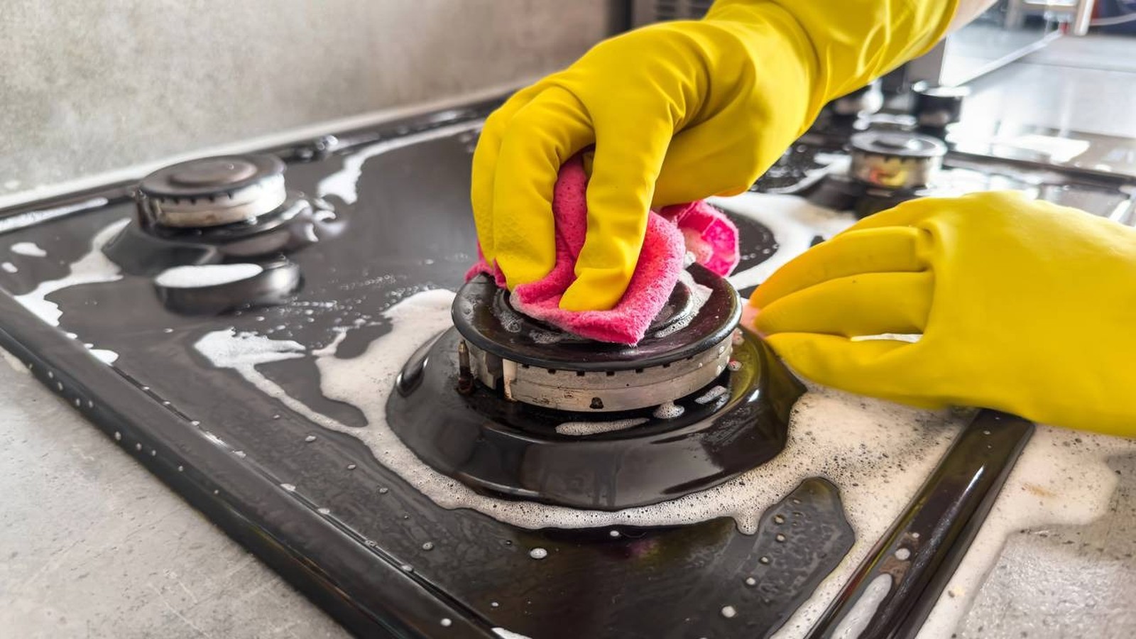 Mãos com luvas amarelas limpam queimador de fogão a gás com pano rosa e espuma
