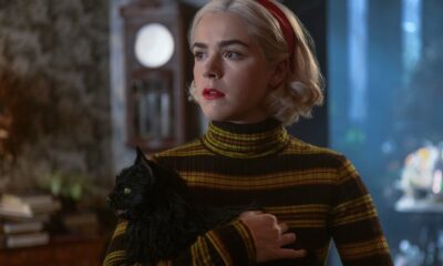 Sabrina Spellman segura um gato preto, usando blusa listrada e bandana vermelha, com olhar sério