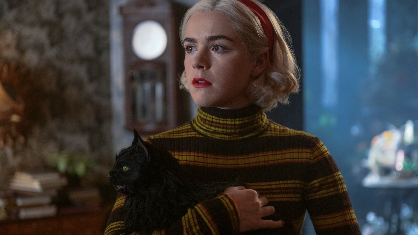 Sabrina Spellman segura um gato preto, usando blusa listrada e bandana vermelha, com olhar sério