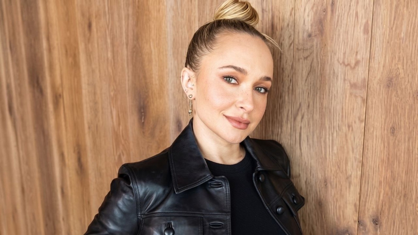 Hayden Panettiere, loira com coque, vestindo jaqueta de couro preta, sorrindo contra fundo de madeira