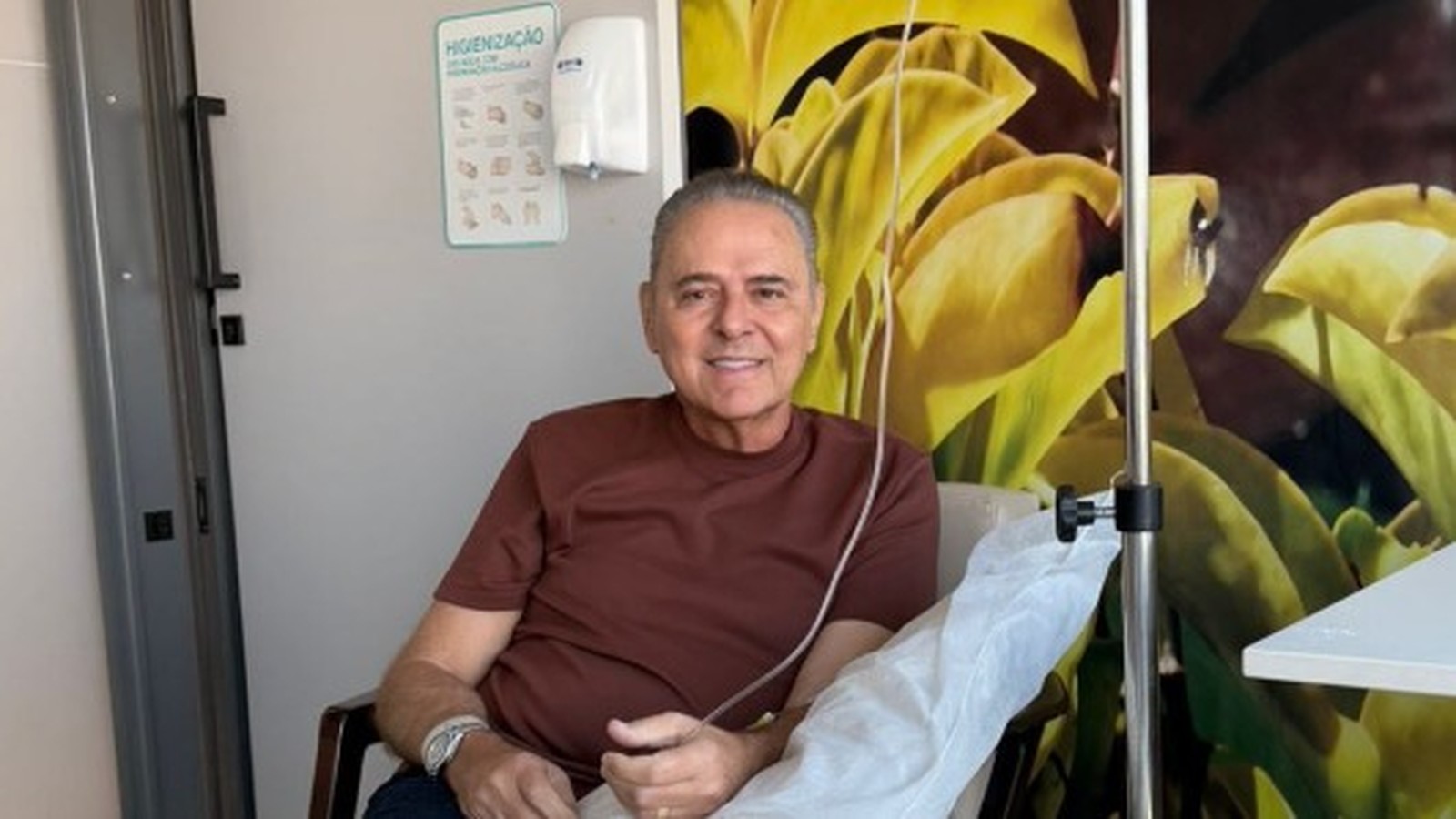 Homem sorrindo com soro intravenoso no braço, sentado em sala com placa de higiene das mãos