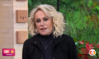 Ana Maria Braga em close, cabelo loiro cacheado, roupa escura. Fundo desfocado com logos