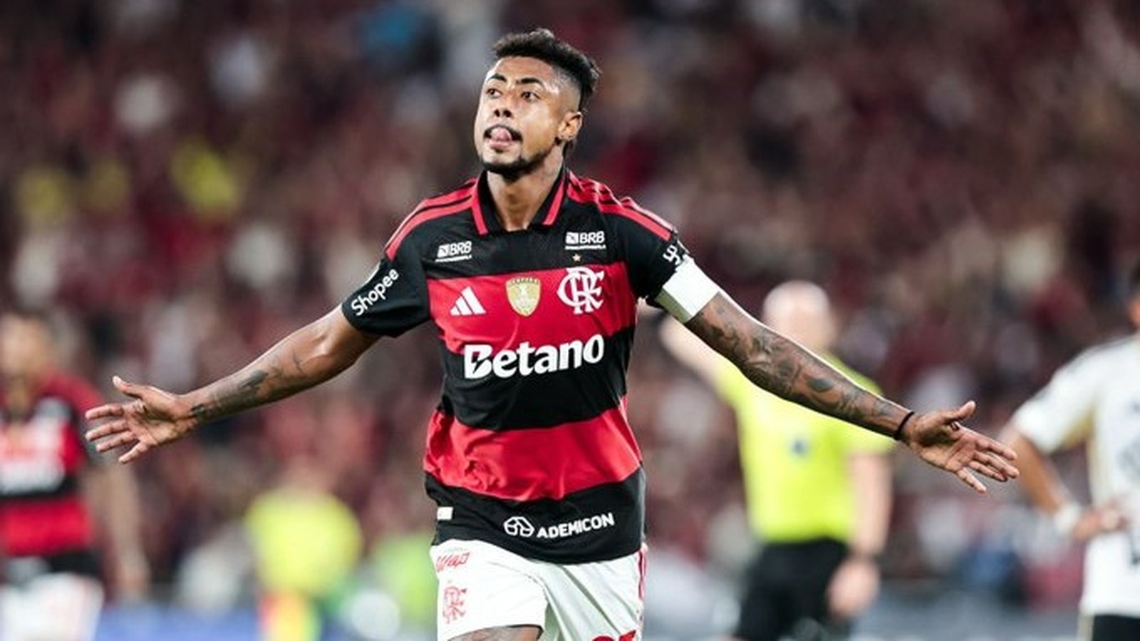 Jogador de futebol Bruno Henrique do Flamengo em camisa vermelha e preta, braços abertos, língua para fora