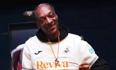 Snoop Dogg sorrindo, vestindo camisa de futebol branca e capuz preto, com colar dourado