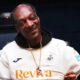 Snoop Dogg sorrindo, vestindo camisa de futebol branca e capuz preto, com colar dourado