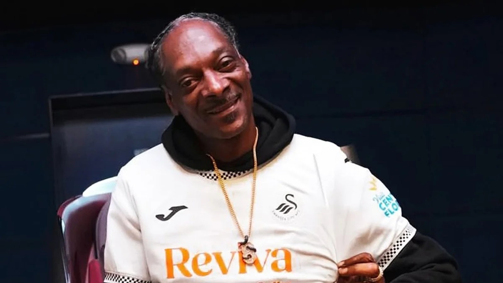 Snoop Dogg sorrindo, vestindo camisa de futebol branca e capuz preto, com colar dourado