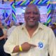 Homem careca sorridente em camisa amarela aponta broche "10" em evento político Republicano
