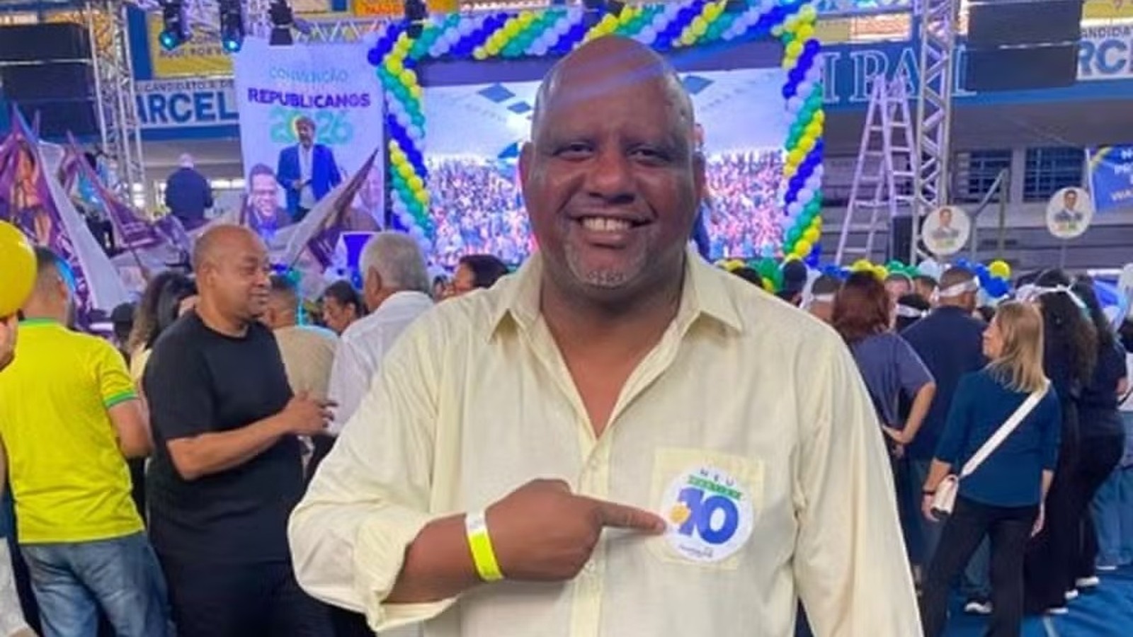 Homem careca sorridente em camisa amarela aponta broche "10" em evento político Republicano