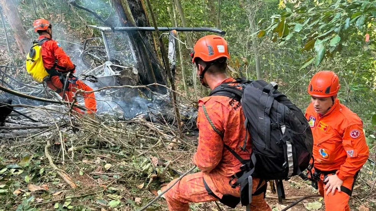 Três bombeiros em floresta densa inspecionam destroços queimados de helicóptero com fumaça
