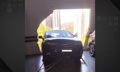 Carro preto atravessando parede de garagem, com edifícios e céu claro ao fundo