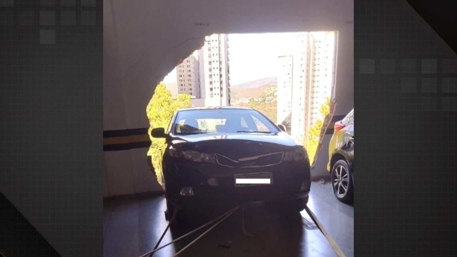 Carro preto atravessando parede de garagem, com edifícios e céu claro ao fundo