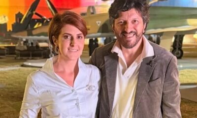 Mila Seidl e Lito Sousa sorrindo juntos, com avião militar ao fundo